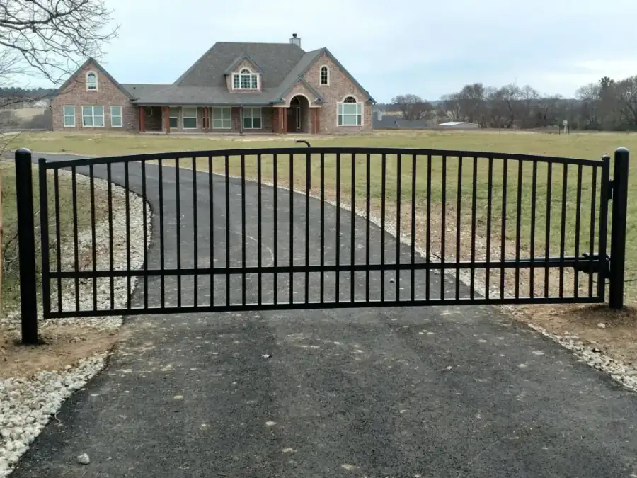 An arched automated wrought iron driveway gate spanning the drive in front of a brick ranch house